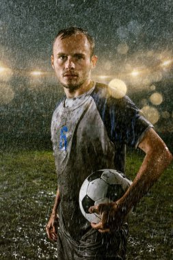 Soccer player on professional soccer night rain stadium. Dirty player in rain drops with football ball clipart