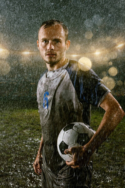 Soccer player on professional soccer night rain stadium. Dirty player in rain drops with football ball