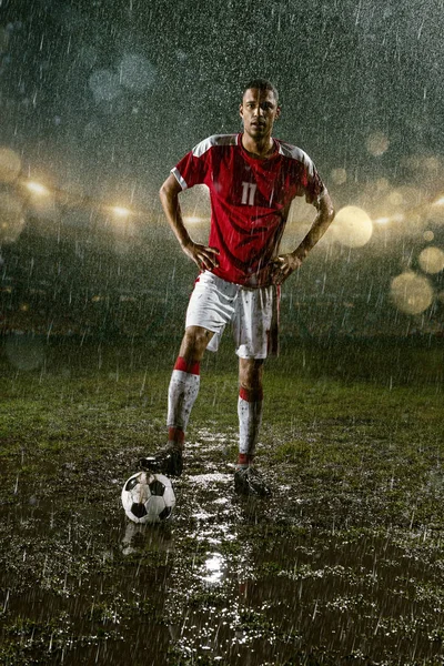 Soccer player on professional soccer night rain stadium. Dirty player in rain drops with football ball