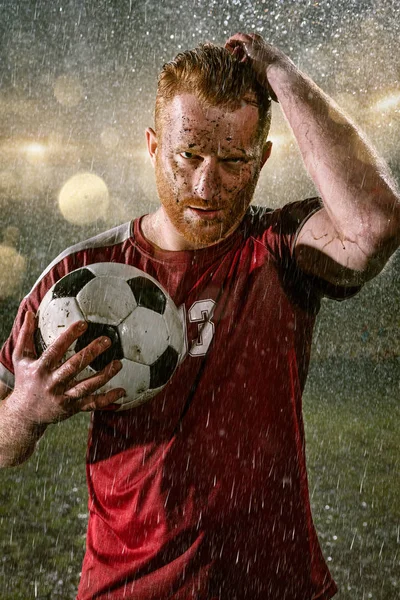 Soccer player on professional soccer night rain stadium. Dirty player in rain drops with football ball