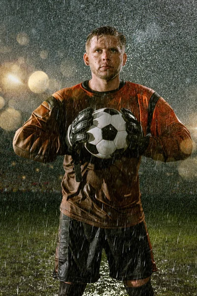 Soccer goalkeeper on professional soccer night rain stadium. Dirty player in rain drops with football ball