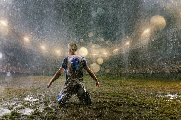 Soccer player on professional soccer night rain stadium. Dirty player in rain drops emotionally rejoices victory. Back view