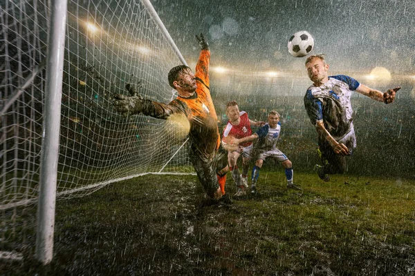 サッカー選手は プロの夜の雨のスタジアムでアクションプレイを行います 雨の中の汚れた選手は 頭でゴールを決めます ボールをキャッチしようとする飛行中のゴールキーパー 雨で濡れたスタジアムの草 — ストック写真