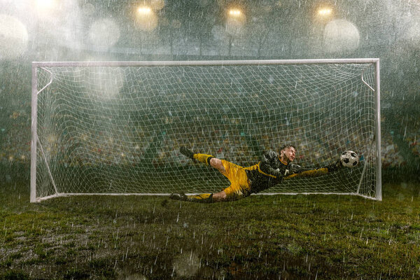 Dirty goalkeeper in flight catch the ball. Professional night rain stadium with football goal. Grass in the stadium wet from the rain