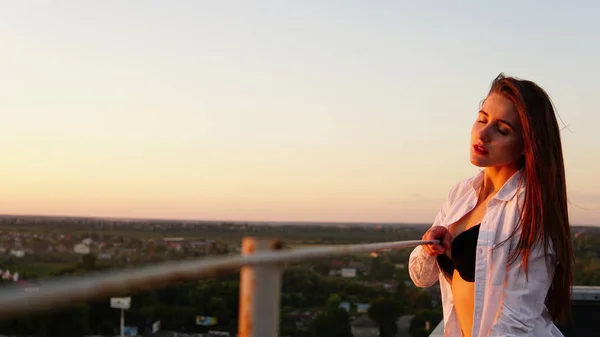 เด็กสาวในชุดกีฬา นั่งอยู่บนขอบหลังคาตอนพระอาทิตย์ตก . — ภาพถ่ายสต็อก