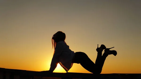 เด็กสาวในชุดกีฬา นั่งอยู่บนขอบหลังคาตอนพระอาทิตย์ตก . — ภาพถ่ายสต็อก