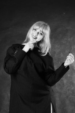 Sandalyeye oturmuş, karanlık stüdyo arka planında poz veren erkek ceketli seksi genç bir kadın. 