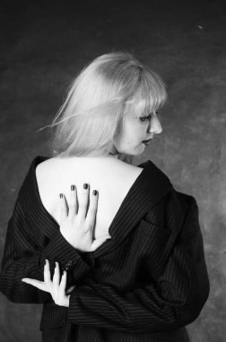 Sandalyeye oturmuş, karanlık stüdyo arka planında poz veren erkek ceketli seksi genç bir kadın. 