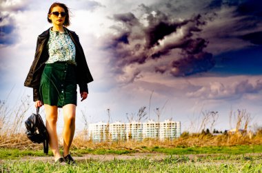 Güzel esmer kadın, arka planda şehir manzarası olan çayırda şık giysiler içinde poz veriyor.