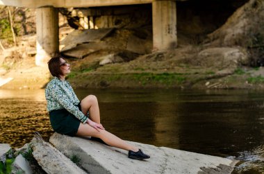 Güneşli bir günde nehir kıyısında şık kıyafetler içinde poz veren güzel esmer kadın.