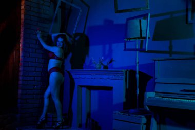 Kırmızı iç çamaşırlı muhteşem kızıl kadın karanlık stüdyoda şöminenin yanında poz veriyor.