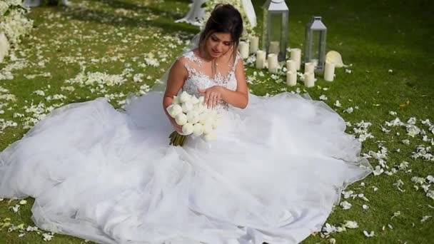 花嫁は緑の芝生の上に結婚式のアーチのそばに座って 彼女の花嫁の花束に触れる — ストック動画