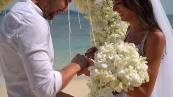 Novia en un vestido blanco, le pone un anillo al novio. arco de flores junto al océano — Vídeo de stock