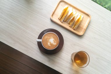 Hafif ahşap bir masada bir tabak sandviçin yanına konmuş sıcak bir fincan kahve. Gündelik yemek için mükemmel ya da lezzetli ısırıkların tadını çıkarmak için rahat anlar..