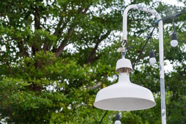 Yeşilliğe karşı sergilenen zarif beyaz lamba, açık hava ayarları için mükemmel, toplanma ve rahatlama için sıcak bir ortam yaratıyor..