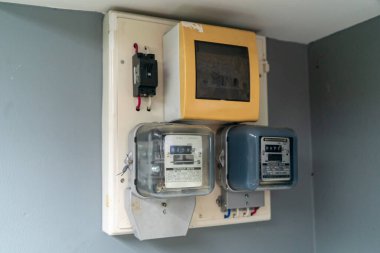 Elektrik sayaçlarının yakın plan görüntüsü ve gri bir duvara monte edilmiş bir devre kesici paneli, konut kullanımı için enerji tüketimi ve ölçüm ekipmanlarını gösteriyor.