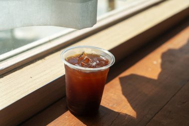 Bu resim, bir cam tarafından yerleştirilmiş bir bardak ferahlatıcı buzlu kahve sergiliyor, sıcak güneş ışığını ve gölgeleri yakalıyor, samimi ve davetkar bir atmosfer yaratıyor..