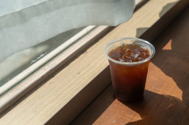 Serinletici bir bardak buzlu kahve, güneşli, ahşap bir masada oturur. Rahat ve davetkar bir anı yakalar. Rahatlamak ya da keyif almak için mükemmel bir an..