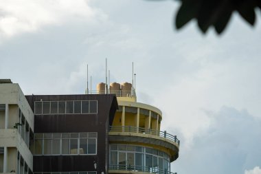 Bu resim, şehir ortamında çağdaş mimari tasarımı vurgulayan, bulutlu bir gökyüzüne karşı belirgin çatı su tanklarının yer aldığı modern bir binayı gösteriyor..