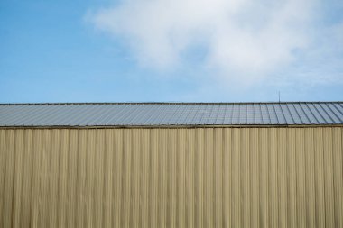 Mimari tasarım ve modern yapı elementlerini ön plana çıkaran, berrak mavi gökyüzüne karşı metal bir çatı sergileyen endüstriyel bir binanın huzurlu manzarası..