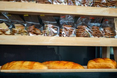 Taze ekmek ve çeşitli hamur işlerinin sergilendiği bir fırın görüntüsü. Ahşap raf altın somunlar ve temiz ambalajlı ikramlar barındırıyor, davetkar bir atmosfer yaratıyor..