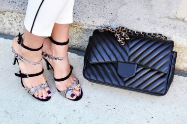 Şık modern moda kadın aksesuar. Beyaz kadın yüksek topuk siyah-yılan derisi bantlı sandalet ve siyah deri çanta beyaz pantolon giyiyor. Ultra violet kadın çivi vardır 