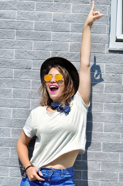 Summer city vibes. Sunny lifestyle fashion portrait pretty young smiling  woman having fun wearing trendy outfit sunglasses with hand raised pointing up over grey background