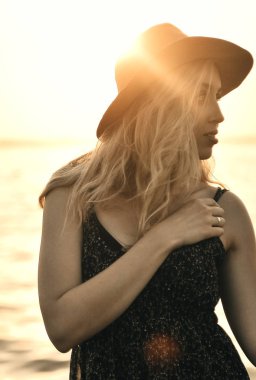 Şapkalı mutlu kadın gün batımında sahilde yürüyor.