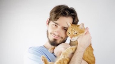 Sakallı bir adam kollarında kırmızı bir kedi tutuyor. Adam mutlu.