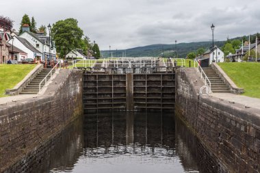 Fort Augustus, İskoçya - 11 Haziran 2012: Kilit Oich Nehri kanalı gösteren yeşil çimenlerin üzerinde kapalı koyu kahverengi kapılar ve ağır gri gökyüzü altında her iki tarafta beyaz evler ile. Ufuk tepelerinde.