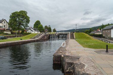 Fort Augustus, İskoçya - 11 Haziran 2012: Kilit Oich Nehri kanalı gösteren yeşil çimenlerin üzerinde kapalı koyu kahverengi kapılar ve ağır gri gökyüzü altında her iki tarafta beyaz evler akan su ile. Ufuk tepelerinde.