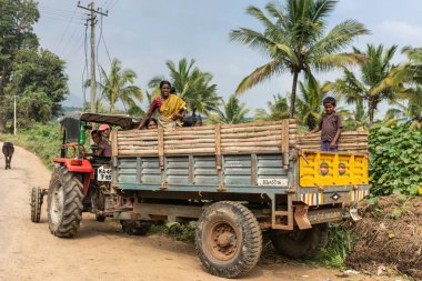 Naralapura, Karnataka, Hindistan - 1 Kasım 2013: Kadın ve çocuk ayağa ve kırmızı traktör üzerinde toprak yol arkada ışık mavi gökyüzü altında yeşil ağaç bitki ile bağladım römork tarım üzerinde oturmak.