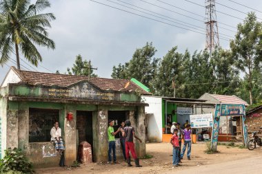 Naralapura, Karnataka, Hindistan - 1 Kasım 2013: Çocuklar, gençler ve yaşlı adam köyde kirli beton toplu taşıma barınağında bekle. Kahve dükkanı bitişik. Yeşil ağaçlar, kahverengi kir ve açık mavi gökyüzü.