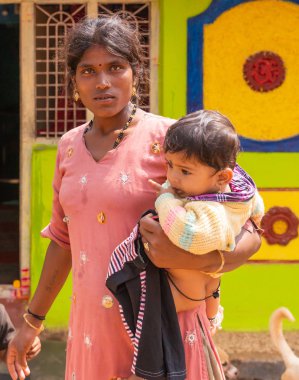 Nilavagilukaval, Karnataka, Hindistan - 1 Kasım 2013: Closeup bakan koyu saçlı Genç Anne kalın giyinik bebek kızı kolundaki pembe elbiseli. Altın takı her iki. Renkli cephe arka plan.
