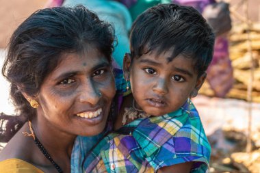 Nilavagilukaval, Karnataka, Hindistan - 1 Kasım 2013: Closeup kolundaki hüzünlü bebek kızı ile gülümseyen koyu saçlı Genç Anne. Altın takı her iki. Çok renkli arka plan soluk.