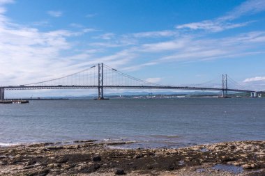 Queensferry, İskoçya, İngiltere - 14 Haziran 2012: İleri yol araçları üzerinde Firth of Forth mavi gökyüzü ve masmavi bir deniz arasında için klasik süspansiyon brige köprüdür. Kayalık sahil ön.