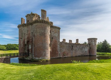 Shearington, İskoçya, İngiltere - 18 Haziran 2012: Giriş ve Batı tarafı yıkık üçgen Brown moated Caerlavarock kale mavi gökyüzü altında taş ve yeşil çim ve orman arkada ayarla.