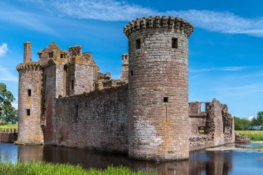 Shearington, İskoçya, İngiltere - 18 Haziran 2012: Güneybatı Kulesi ve harap kahverengi taş Castle Caerlavarock mavi gökyüzü altında iki duvar. Geniş hendek, yeşil çim ve ağaçların.