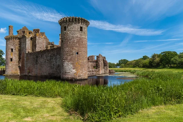 Shearington, İskoçya, İngiltere - 18 Haziran 2012: Güneybatı Kulesi ve harap kahverengi taş Castle Caerlavarock mavi gökyüzü altında iki duvar düşük ihtimal. Geniş hendek, yeşil çim ve ağaçların.