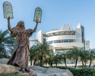 Garden Grove, California, USA - December 13, 2018: Crystal Christ Cathedral. Bronze statue of Moses putting the ten commandments of two tables in the air. Some green foliage, blue sky, white Cultural Center in back. clipart