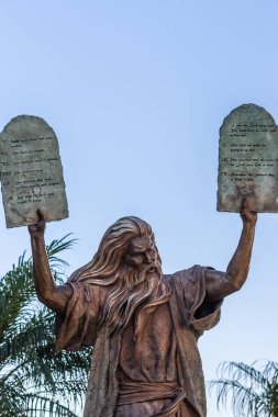 Garden Grove, California, USA - December 13, 2018: Crystal Christ Cathedral. Closeup of Bronze statue of Moses putting the ten commandments of two tables in the air. Some green foliage, light blue sky. clipart