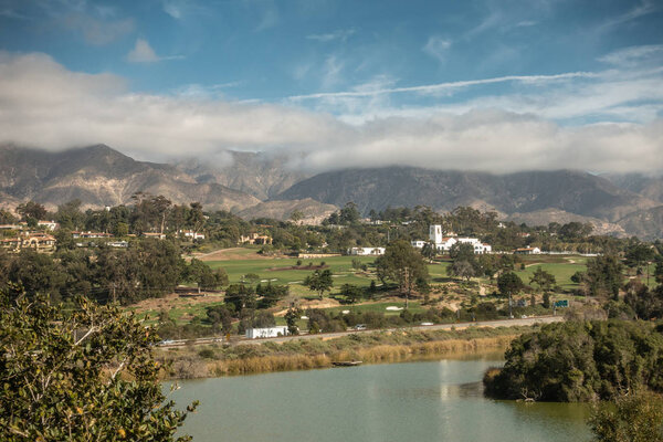 Santa Barbara, California, USA - December 18, 2018: Green golf course and white towered building of Montecito Country Club. Mountains partly shrouded in clouds under blue sky in back. Bird Refuge waters in front.