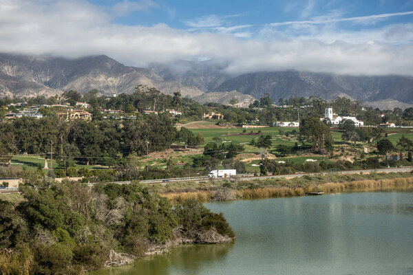 Santa Barbara, California, USA - December 18, 2018: Green golf course and white towered building of Montecito Country Club. Mountains partly shrouded in clouds under blue sky in back. Bird Refuge waters in front.