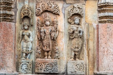 Belur, Karnataka, India - November 2, 2013: Brown wall stone panel sculpture of Lord Vishnu surrouned by his two consorts Devi Lakshmi and Devi Bhu with Shilabalika women. On side of temple building. clipart