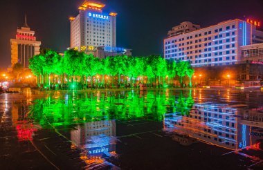 Xian, Çin - 1 Mayıs 2010: Kara gece manzarası. Şehir merkezindeki HNA Oteli ve yeşil ışıklı ağaçlar ve ıslak yüzey yansımaları parkının üzerinde görülen diğer yüksek binalar aydınlandı..