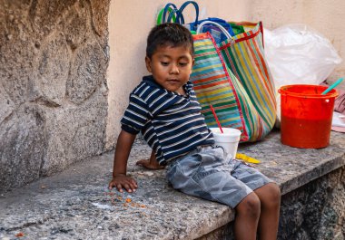 Acapulco, Meksika - 25 Kasım 2008: Old Downtown. Mavi gömlekli ve gri şortlu genç adam gri taş çıkıntıda oturuyor ve bekliyor. Arkasında da kan kırmızısı kovası ve Coloful alışveriş torbaları..