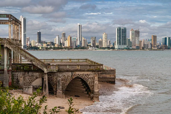Panama City, Panama - November 30, 2008: Modern highrise building along gray bay water shoreline seen from historic downtown under blue cloudscape. Old lookout point in brown stones up front.