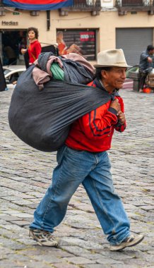Quito, Ekvador - 2 Aralık 2008: Tarihi Şehir Merkezi. Kırmızı gömlekli ve sırtında kıyafetlerle meydanda yürüyen büyük siyah çantalı kot pantolonlu bir adam. Arka tarafta sokak sahnesi.