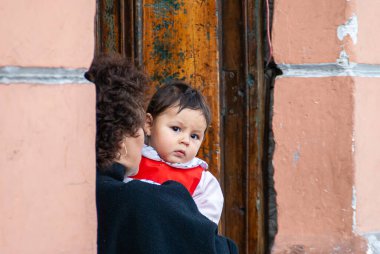 Quito, Ekvador - 2 Aralık 2008: Şehir merkezi tarihi. Mavili annenin kollarında, beyaz ve kırmızı elbiseli küçük kızın yakın çekimi. Pembe taşlı kahverengi kapı..