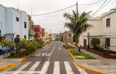 Lima, Peru - 4 Aralık 2008: Gümüş gökyüzü altında daha iyi bir muhitte tarafları yeşil olan boş caddede basit evler. Evler gri, yeşil, bej ve kırmızıya boyanmıştır. Güneş çiçekleri renk ekler.